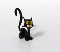 May include: A black glass figurine of a cat with yellow eyes. The cat has a long, curved tail and sits upright with its front legs together.  The figurine is a whimsical, cartoonish representation of a black cat, suitable for home decor or as a collectible. The glass is shiny and reflects light, giving it a glossy appearance. The cat's features are exaggerated, with large, expressive eyes and a slightly mischievous expression.