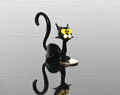 May include: Miniature black glass cat figurine with yellow eyes. The cat is sitting with its tail curled. The figurine is on a dark reflective surface with a coin next to it.  The glass cat is a whimsical, handcrafted piece, perfect for collectors of unique glass art or animal figurines.