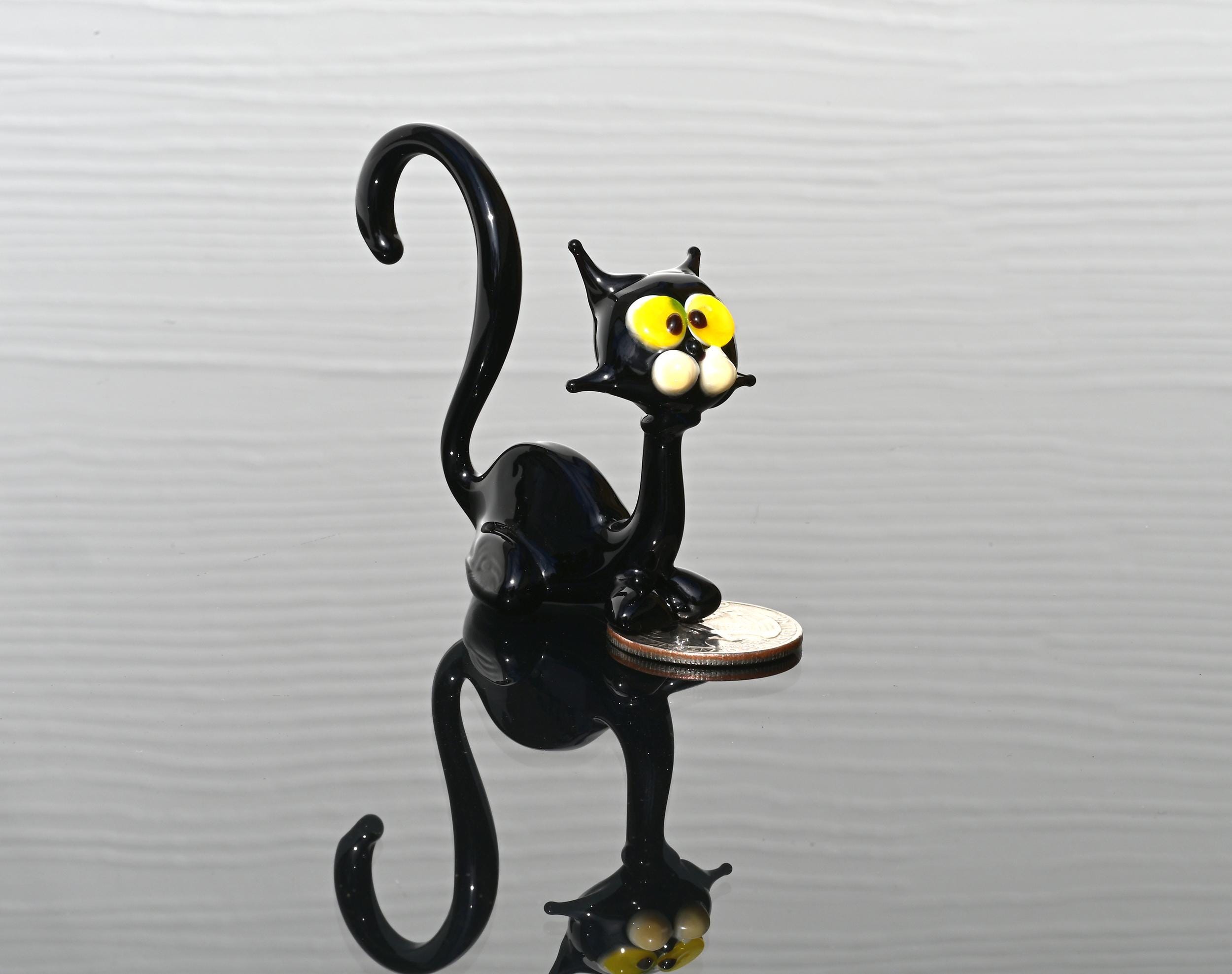 May include: Miniature black glass cat figurine with yellow eyes. The cat is sitting with its tail curled. The figurine is on a dark reflective surface with a coin next to it.  The glass cat is a whimsical, handcrafted piece, perfect for collectors of unique glass art or animal figurines.