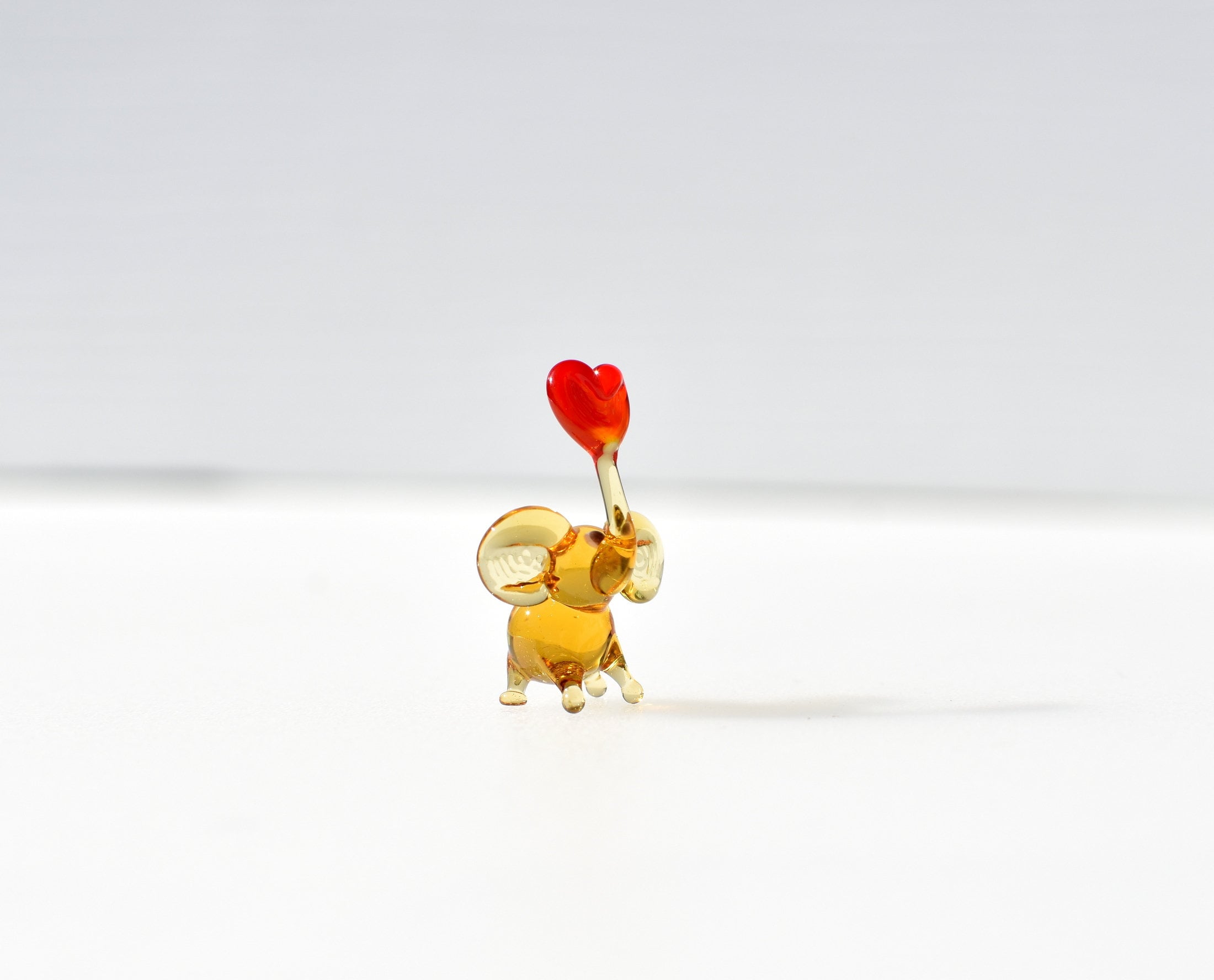 May include: A small glass elephant figurine with a red heart in its trunk. The elephant is a light amber color and is standing on a white surface.