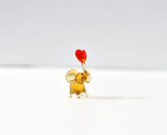 May include: A small glass elephant figurine with a red heart in its trunk. The elephant is a light amber color and is standing on a white surface.