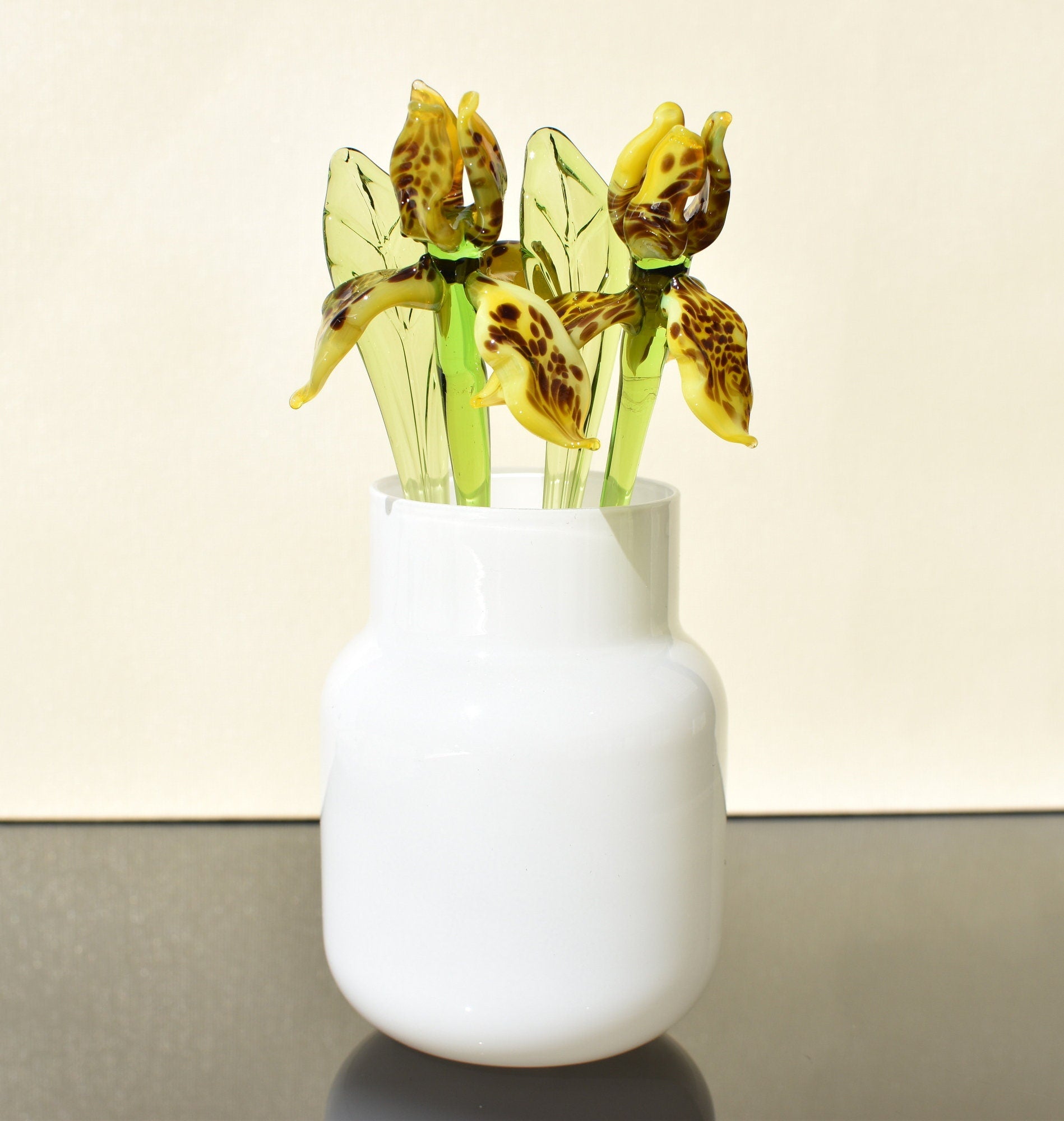 May include: Three glass flowers in a white vase. The flowers are yellow with brown spots and have green stems and leaves.