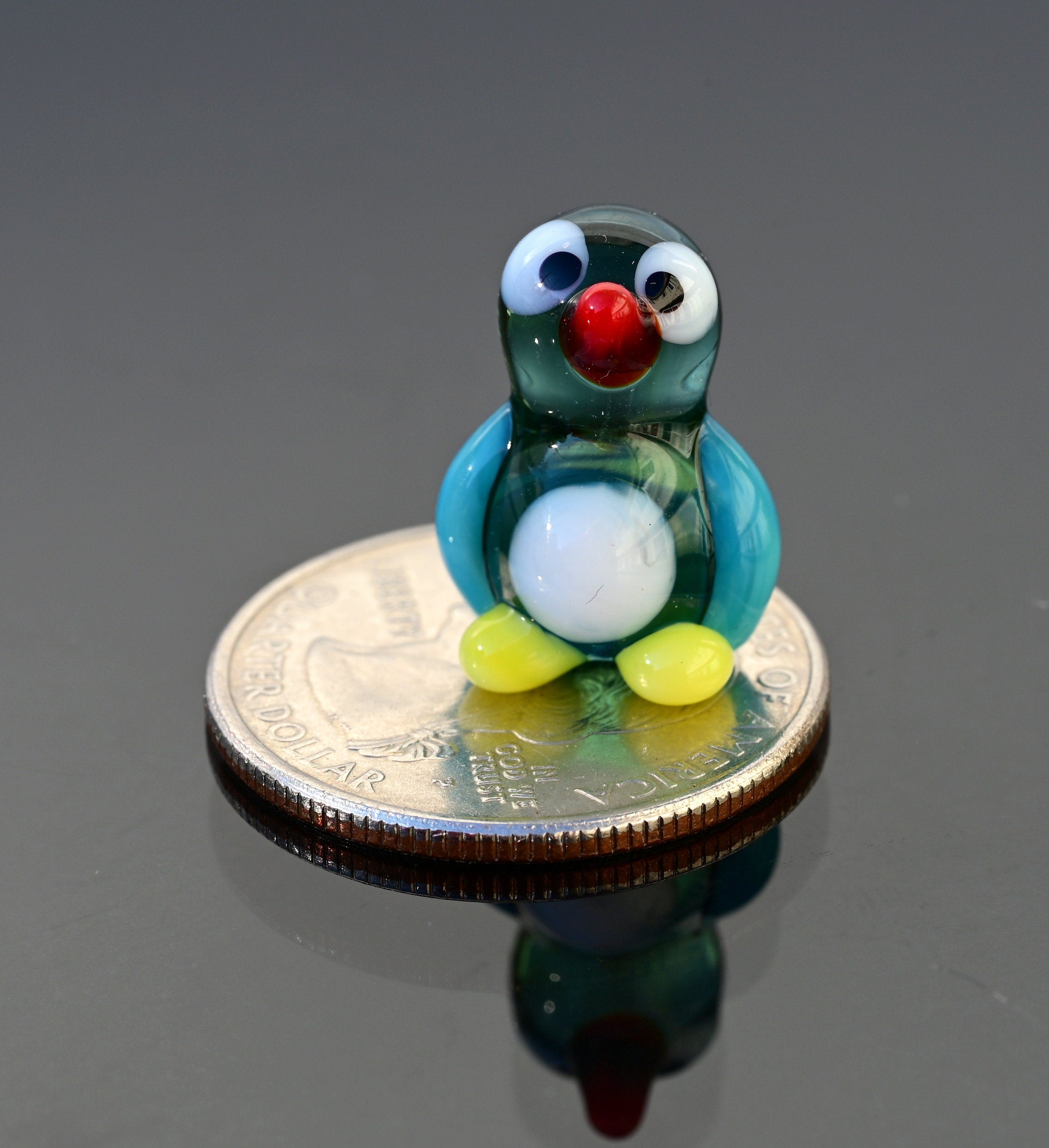 May include: A small glass penguin figurine with a blue and green body, white belly, red beak, and black eyes. The figurine is sitting on a silver US quarter dollar coin.