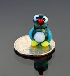 May include: A small glass penguin figurine with a blue and green body, white belly, red beak, and black eyes. The figurine is sitting on a silver US quarter dollar coin.
