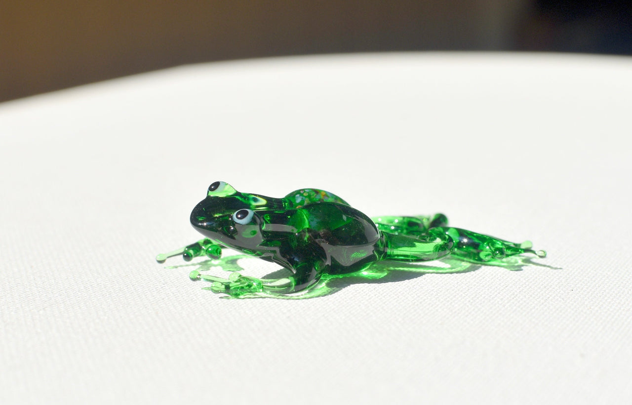 May include: A green glass frog figurine with black eyes. The frog is posed in a sitting position with its legs outstretched.