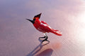May include: A red glass bird figurine with black legs and a black beak. The bird is standing on a light pink surface.