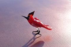 May include: A red glass bird figurine with black legs and a black beak. The bird is standing on a light pink surface.