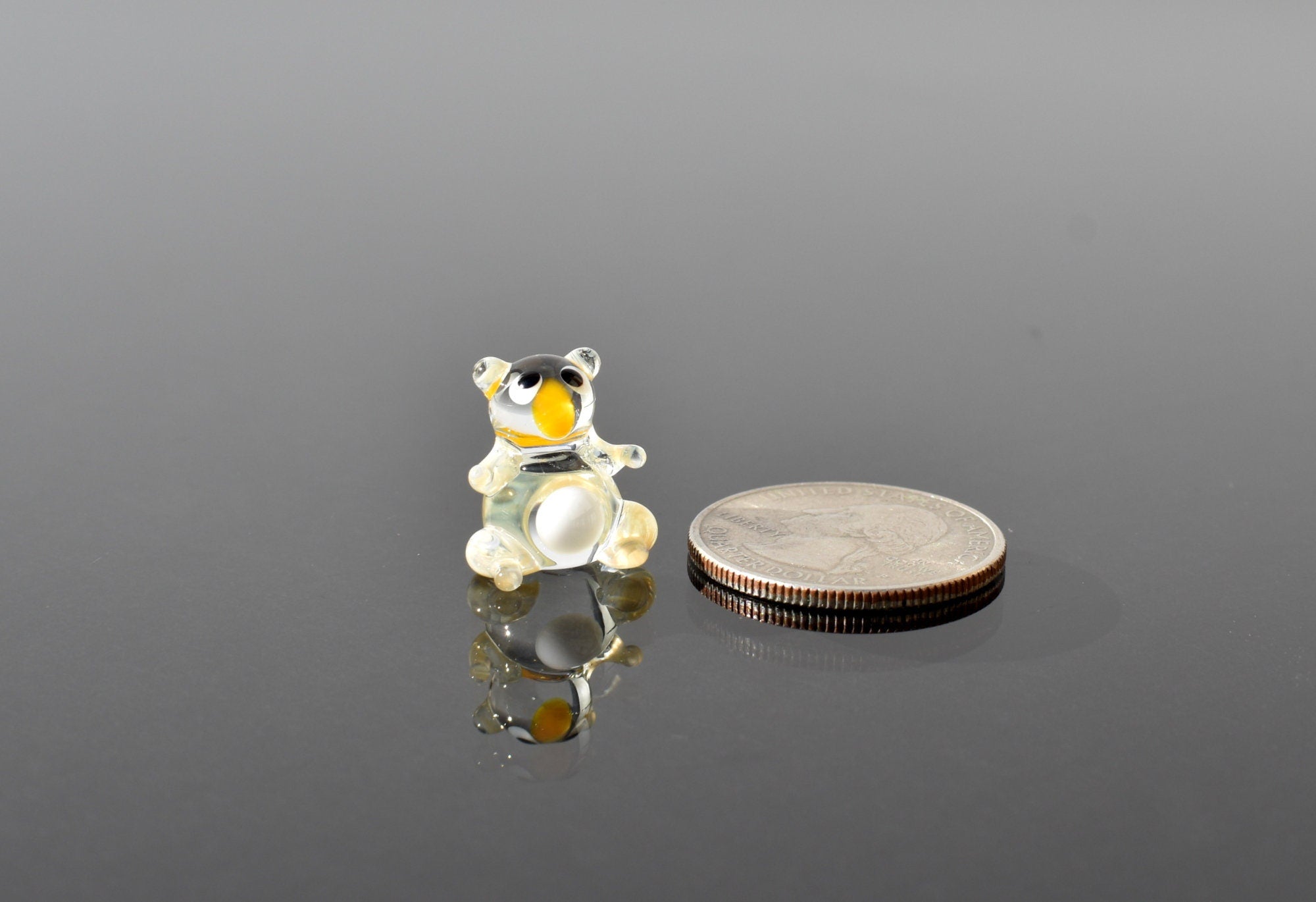 May include: A small, clear glass bear figurine with black eyes and a yellow nose. The figurine is sitting on a reflective surface and is next to a silver US quarter.