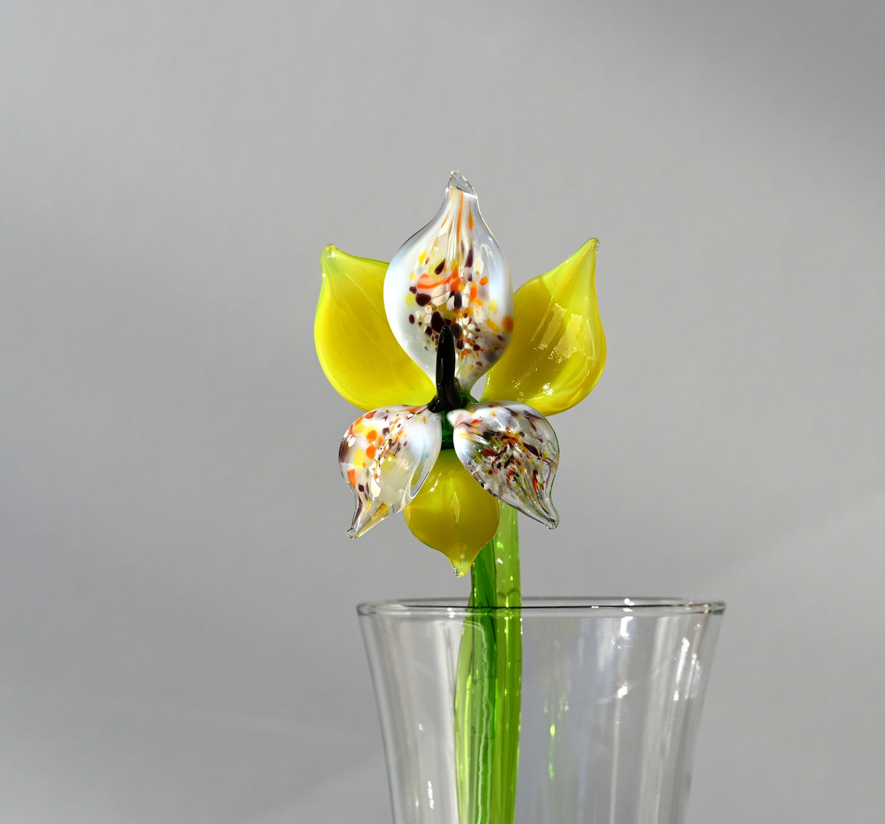 May include: A yellow glass flower with white and orange accents in a clear glass vase. The flower has a green stem and is a realistic representation of a flower.