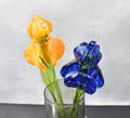 May include: Two glass flowers in a clear glass vase. One flower is orange and the other is blue. The flowers are arranged in a vase on a black surface.