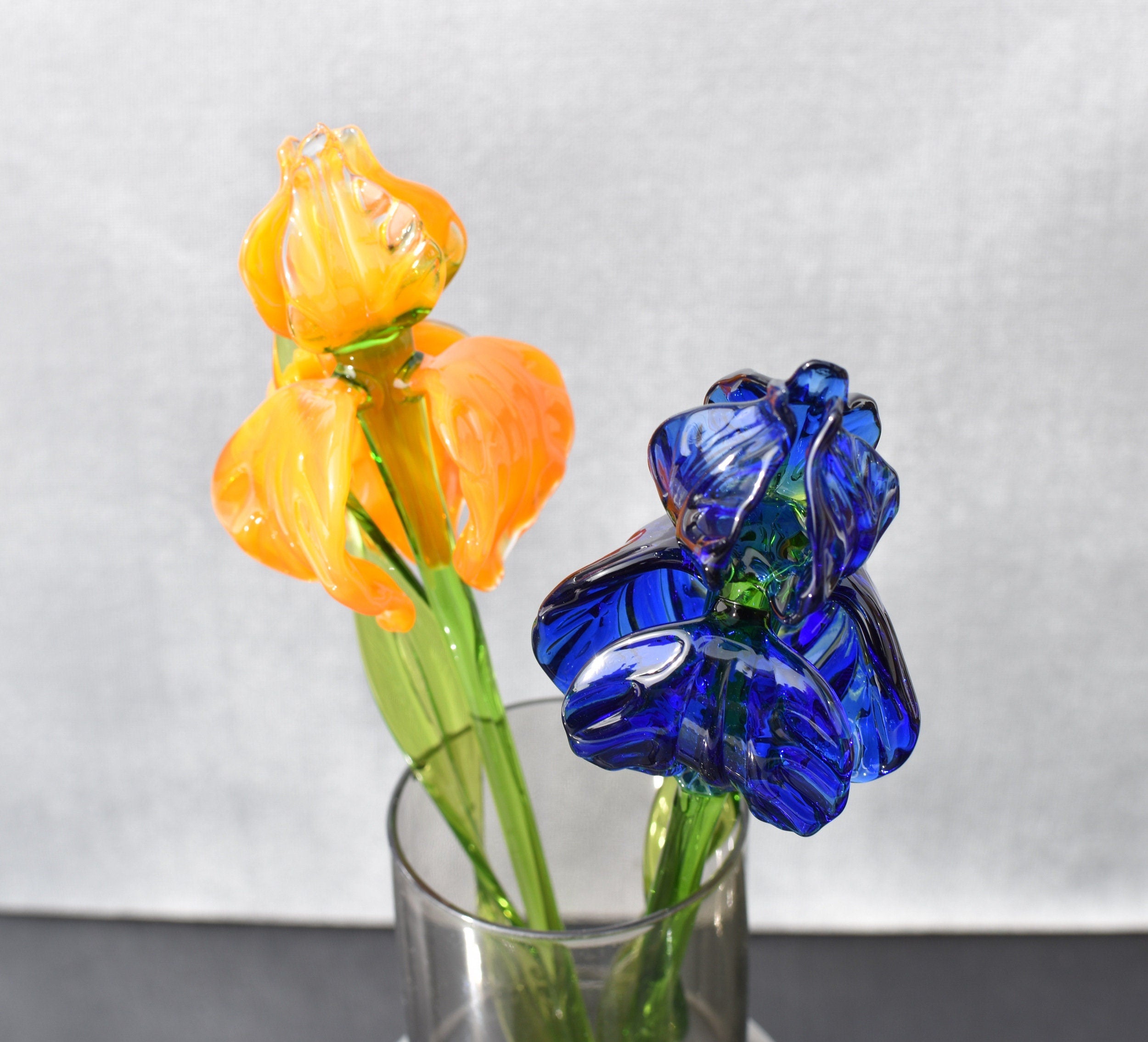 May include: Two glass flowers in a clear glass vase. One flower is orange and the other is blue. The flowers are arranged in a vase on a black surface.