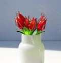 May include: A bouquet of red glass tulips in a white vase. The tulips are made of clear glass with red coloring inside. The vase is a simple, white, round shape.