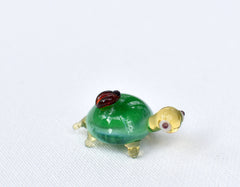 May include: A small glass turtle figurine with a green shell, yellow head, and red eyes. The turtle is sitting on a white surface.