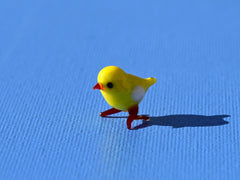May include: A small yellow glass figurine of a chick with red legs and a white spot on its chest. The chick is standing on a blue surface.