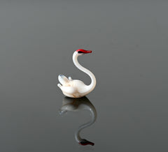 May include: A small white glass swan figurine with a red beak. The swan is in a swimming pose with its head turned to the side.