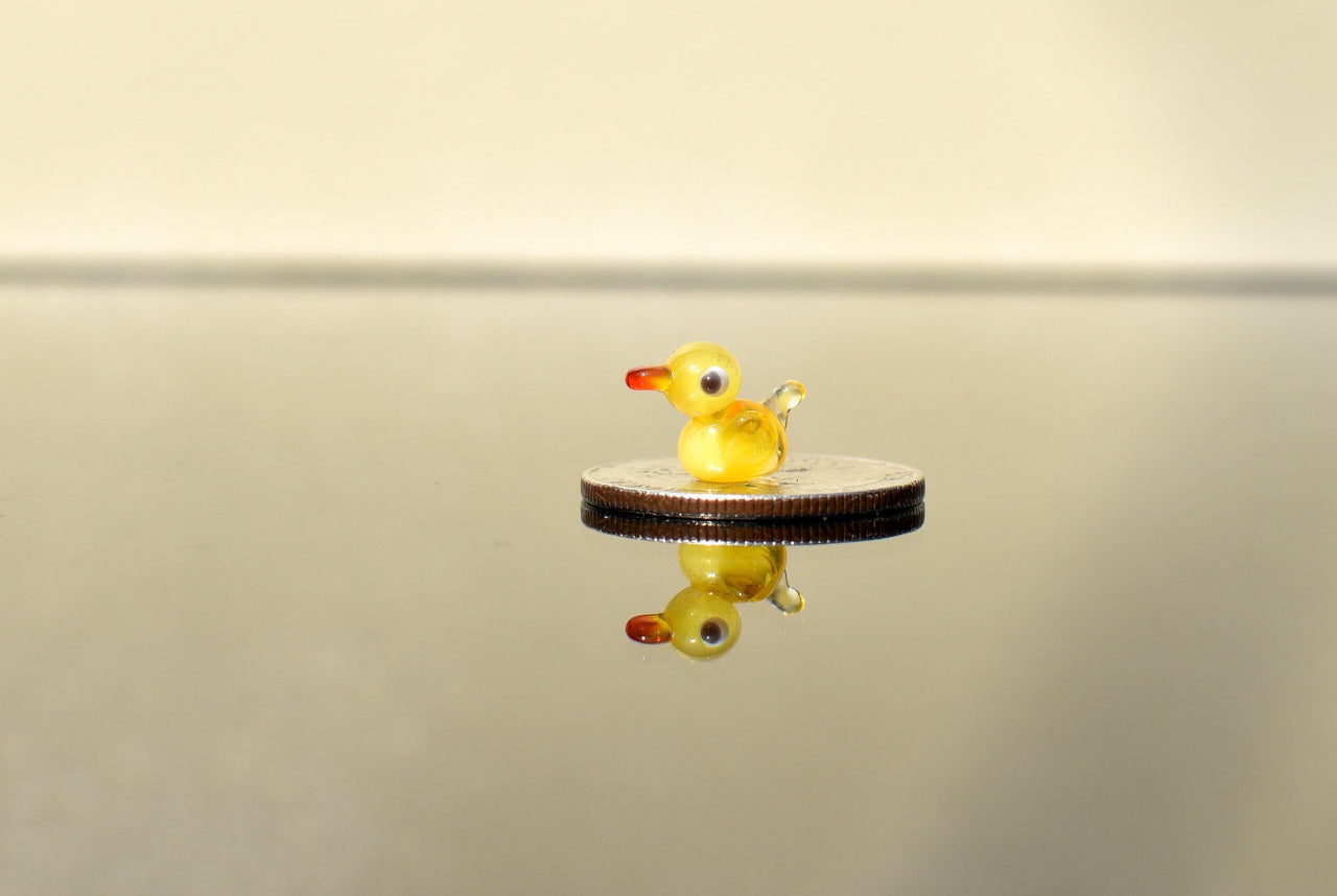 May include: A small yellow glass duck figurine with red eyes and a red beak sitting on a silver coin.