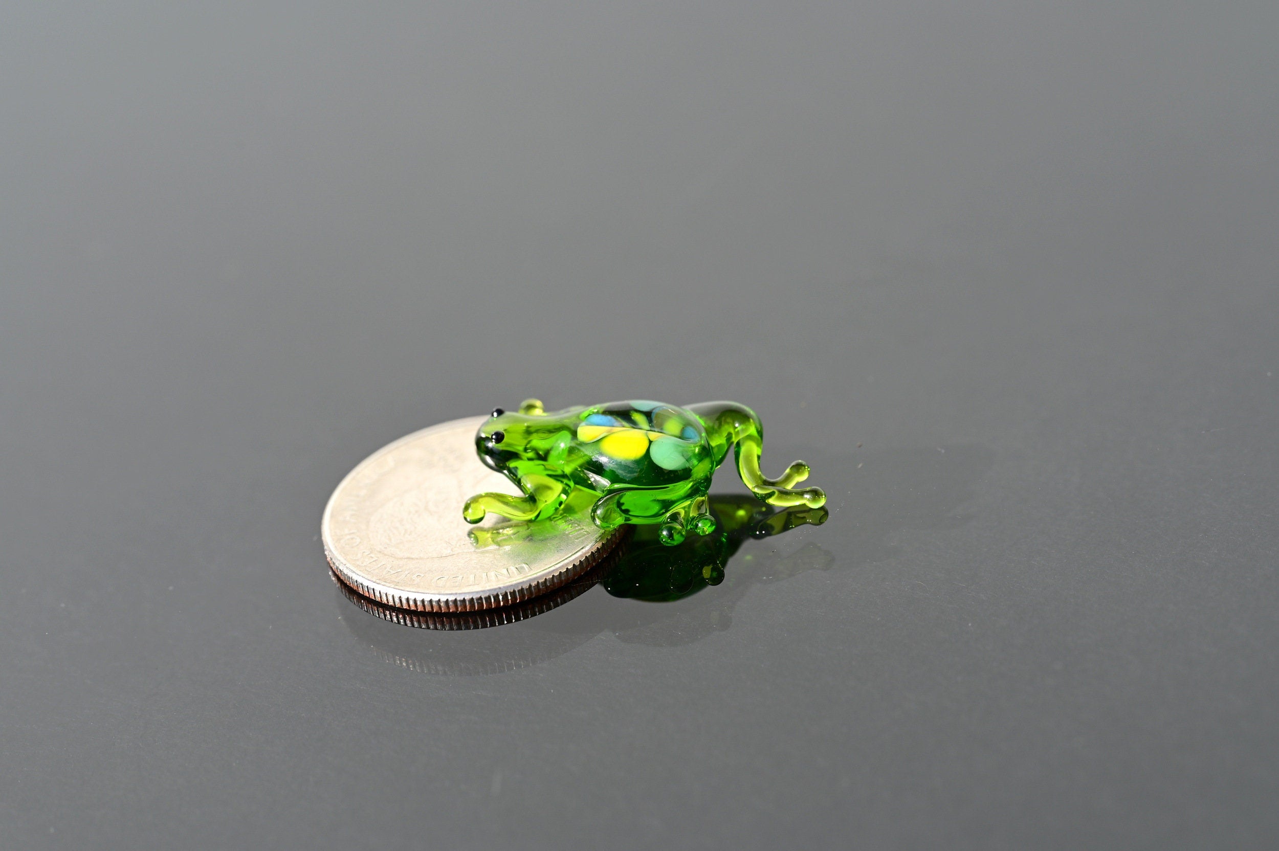 May include: A small green glass frog figurine with yellow and blue accents. The frog is sitting on a silver coin.