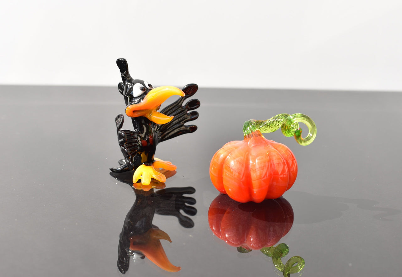 May include: A black glass crow figurine with orange beak and feet, looking up at a small orange glass pumpkin figurine with a green stem.
