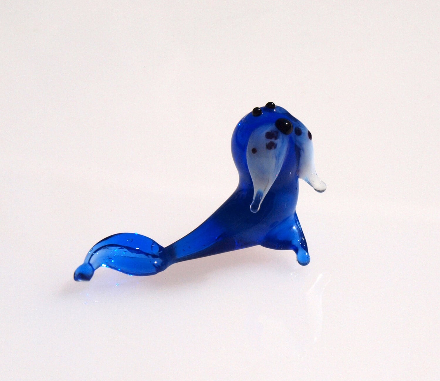 May include: A small blue glass figurine of a seal with white and black details. The seal is lying on its belly with its flippers outstretched.