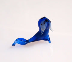 May include: A small blue glass figurine of a seal with white and black details. The seal is lying on its belly with its flippers outstretched.
