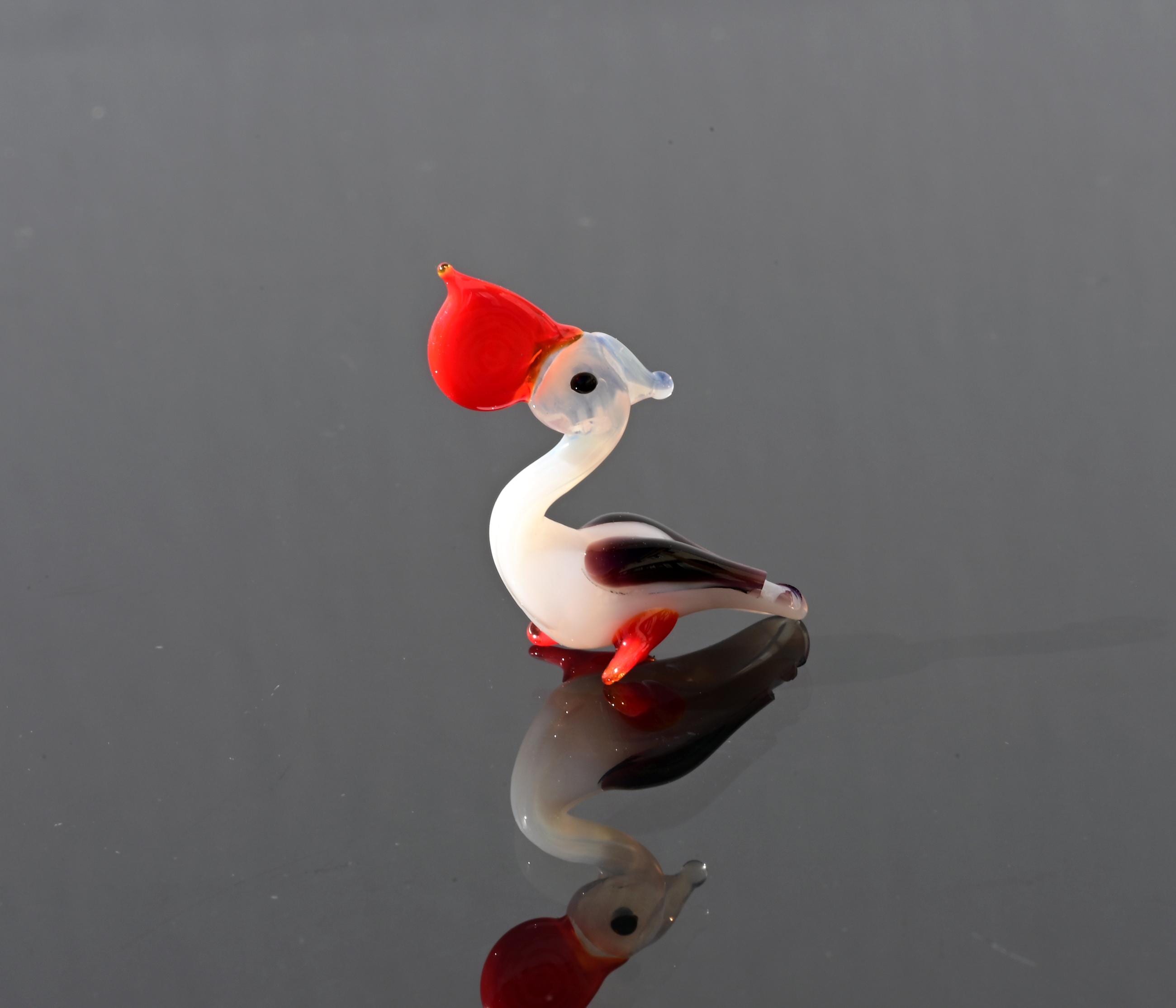 May include: A small glass figurine of a bird with a red head, white body, and black and white wings. The bird is standing on a reflective surface.