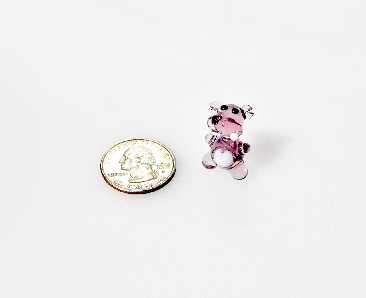 May include: A small glass bear figurine with a purple body and white accents. The bear is standing on a white surface.