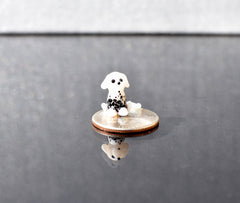 May include: A small, white glass figurine of a dalmatian dog sitting on a silver US quarter. The dog has black spots and is looking to the right. The figurine is sitting on a black surface.