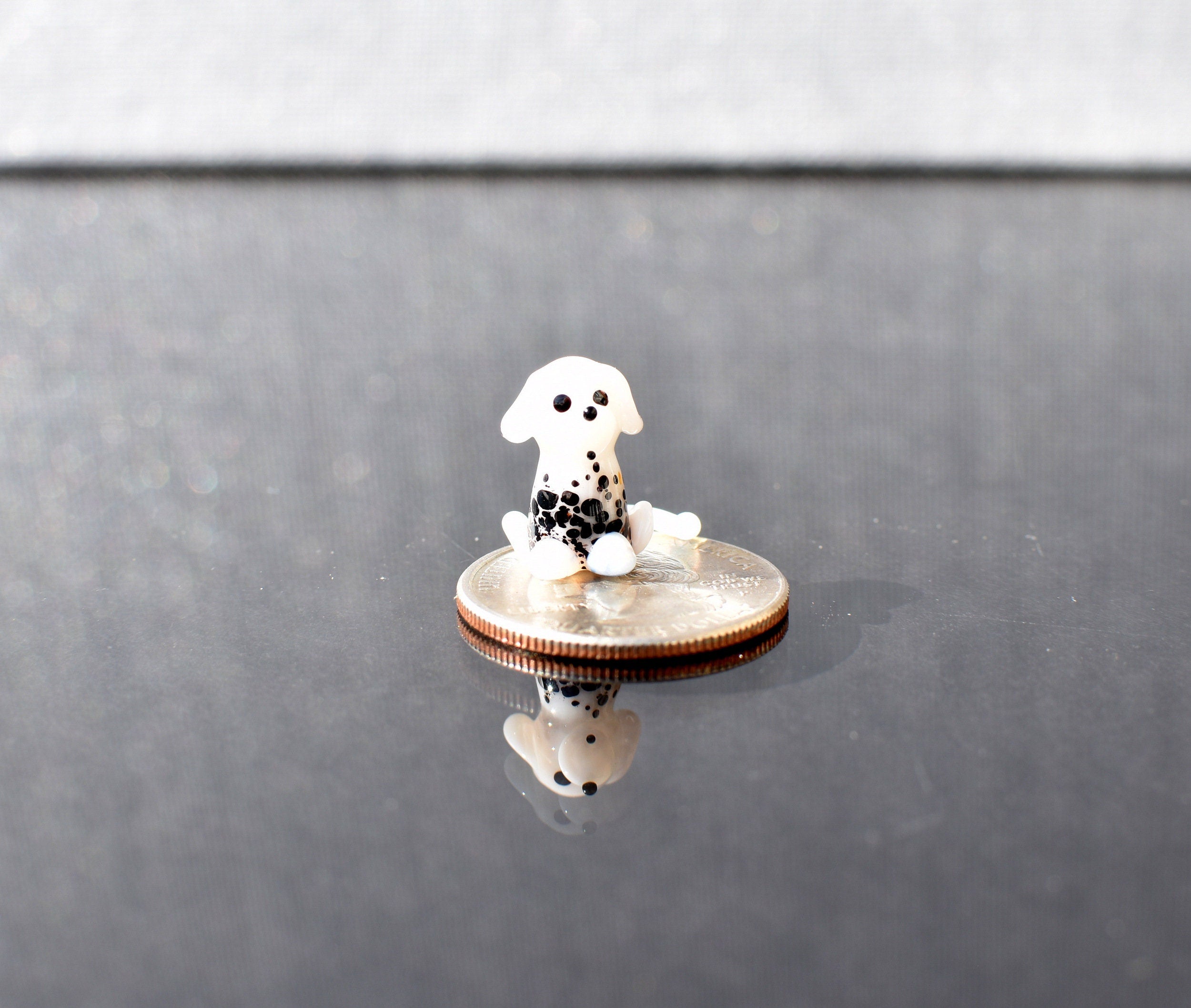 May include: A small, white glass figurine of a dalmatian dog sitting on a silver US quarter. The dog has black spots and is looking to the right. The figurine is sitting on a black surface.