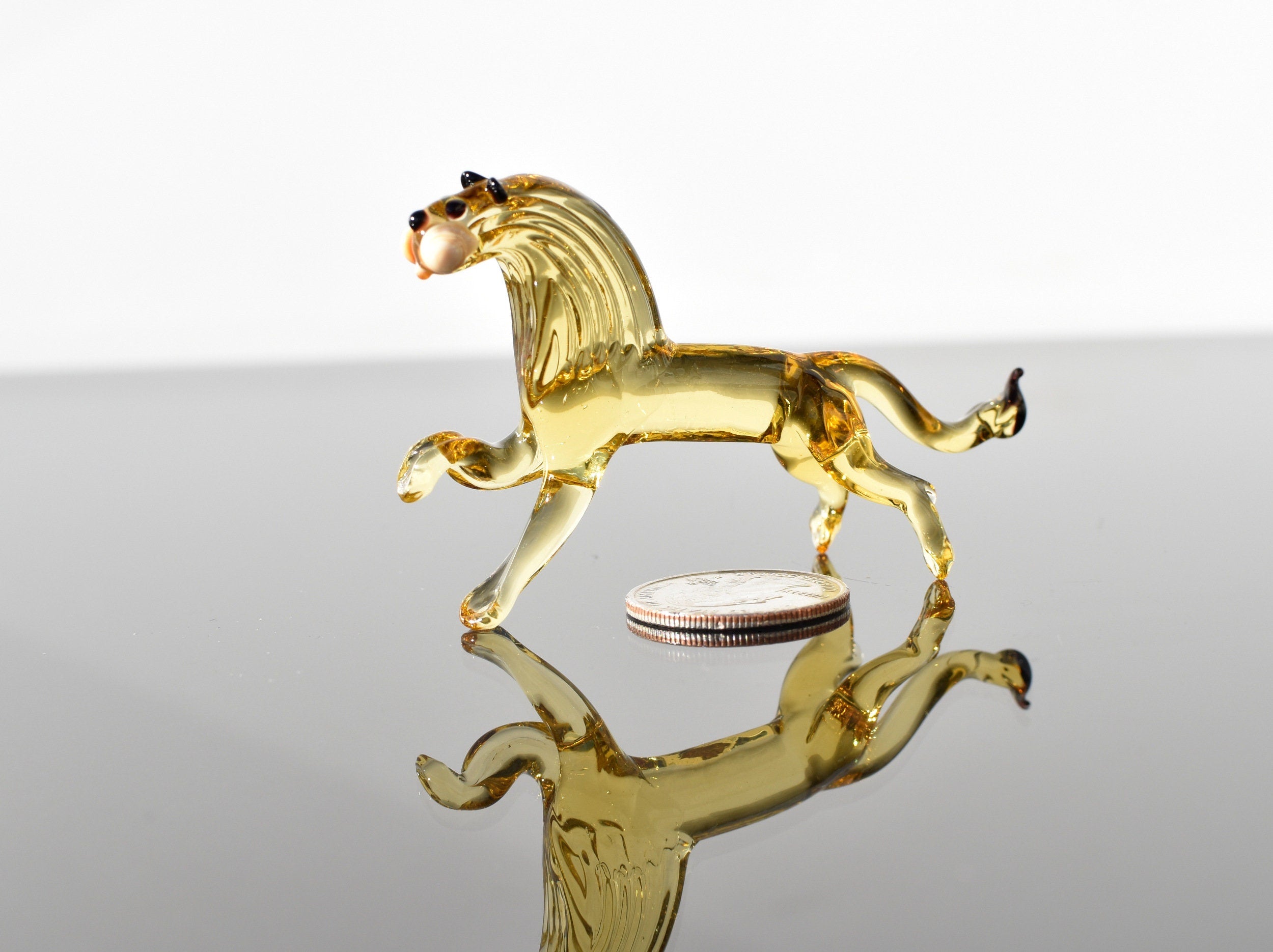 May include: A small glass lion figurine in a yellow amber color. The figurine is standing on a reflective surface with a silver coin in the background.