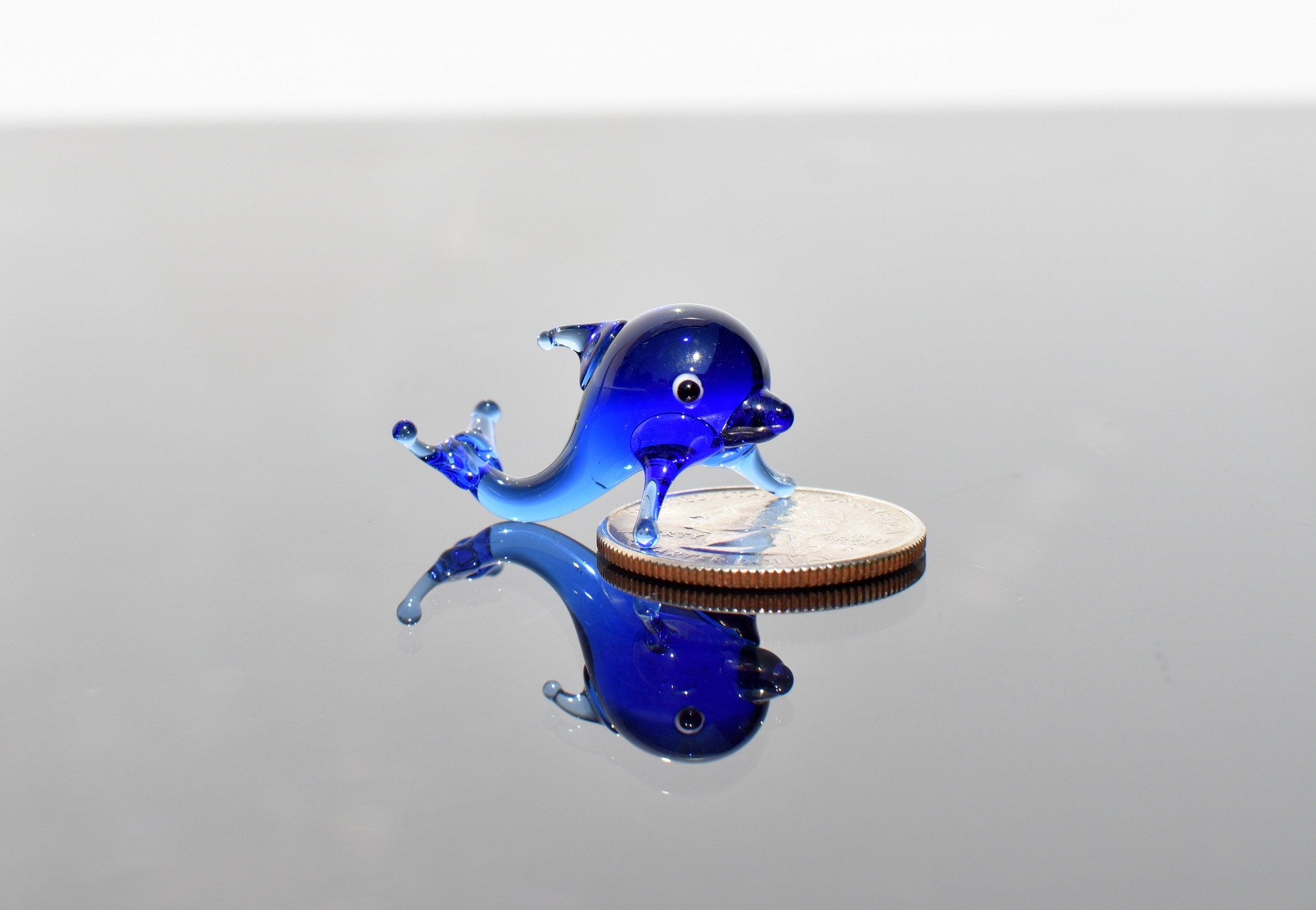 May include: A small blue glass dolphin figurine on a silver coin. The dolphin is facing right and has a white eye. The dolphin is about the size of a quarter.