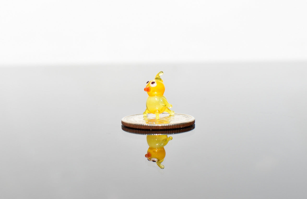 May include: A small yellow glass bird figurine with orange eyes and beak, standing on a silver coin.
