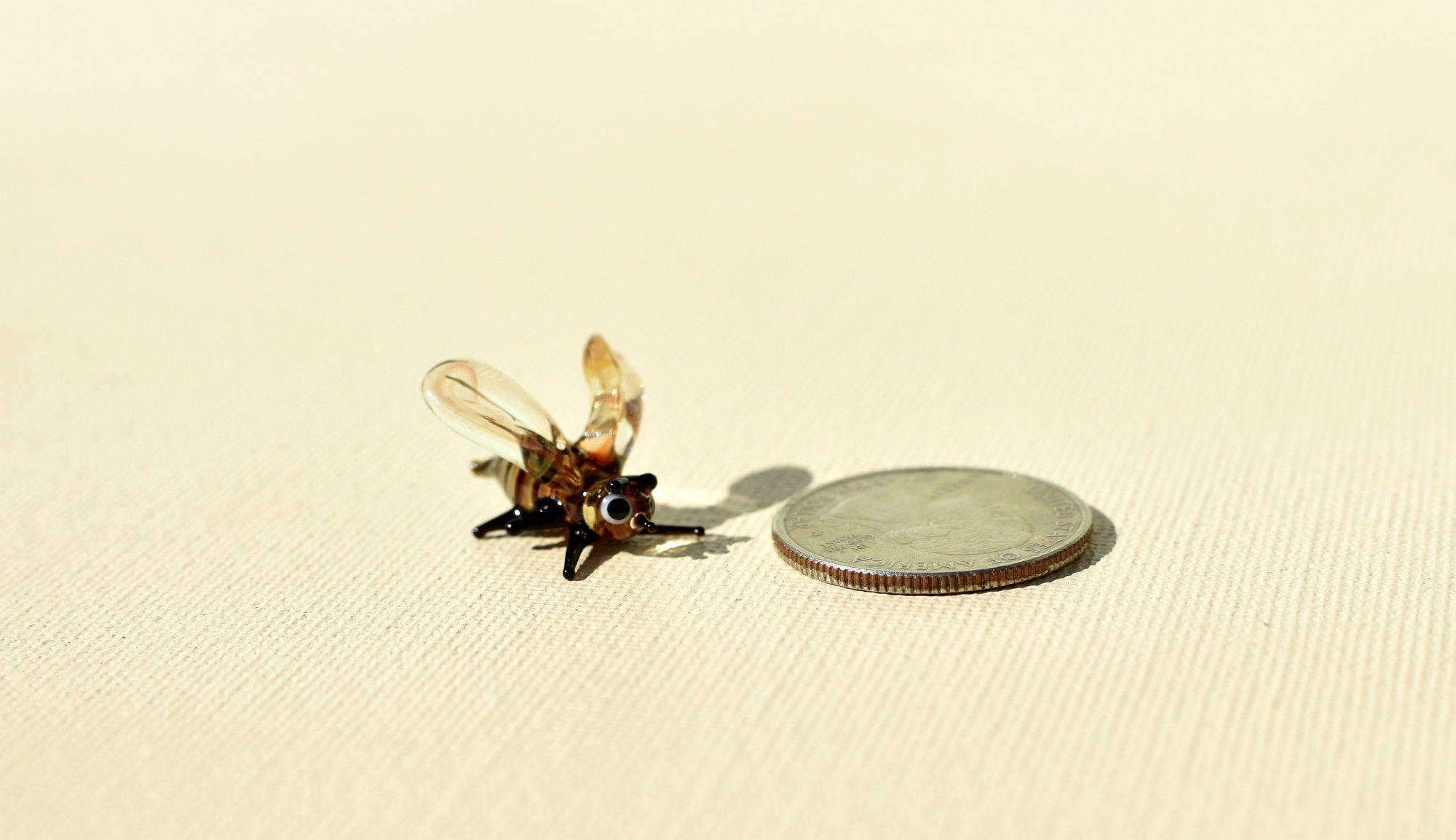 May include: A small glass bee figurine with brown and clear wings. The figurine is sitting on a beige surface next to a silver US quarter coin.