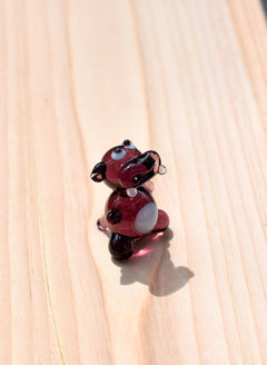 May include: A small glass figurine of a purple hippopotamus with white eyes and a white spot on its belly.