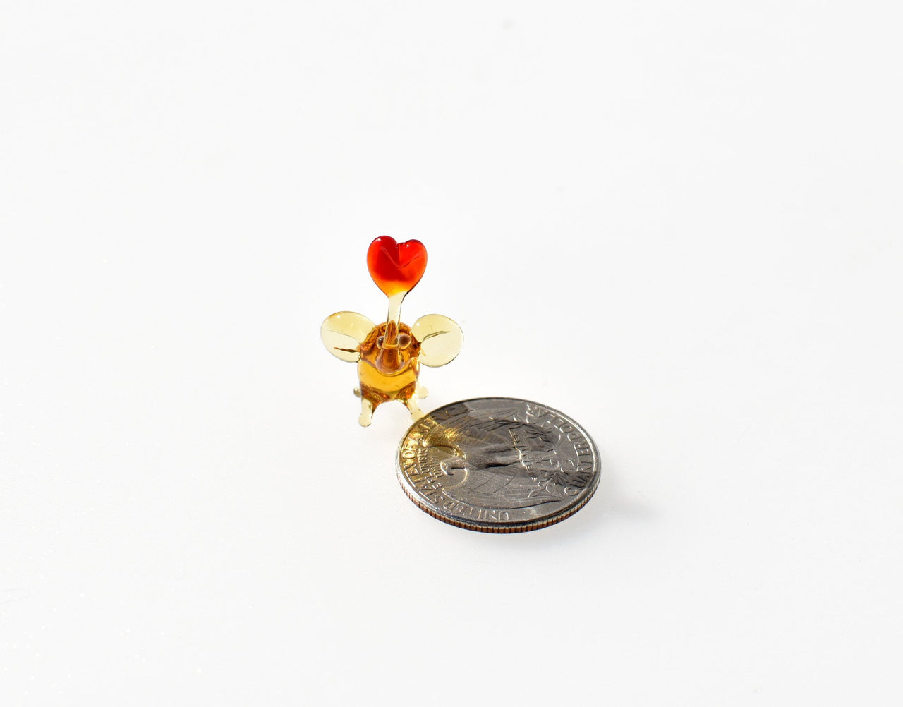 May include: A small glass figurine of a mouse with a red heart on top. The mouse is amber colored and has wings. The figurine is next to a US quarter for size comparison.