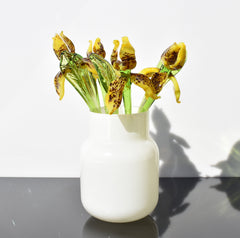 May include: A white ceramic vase with a bouquet of glass flowers. The flowers are yellow with brown spots and green stems.