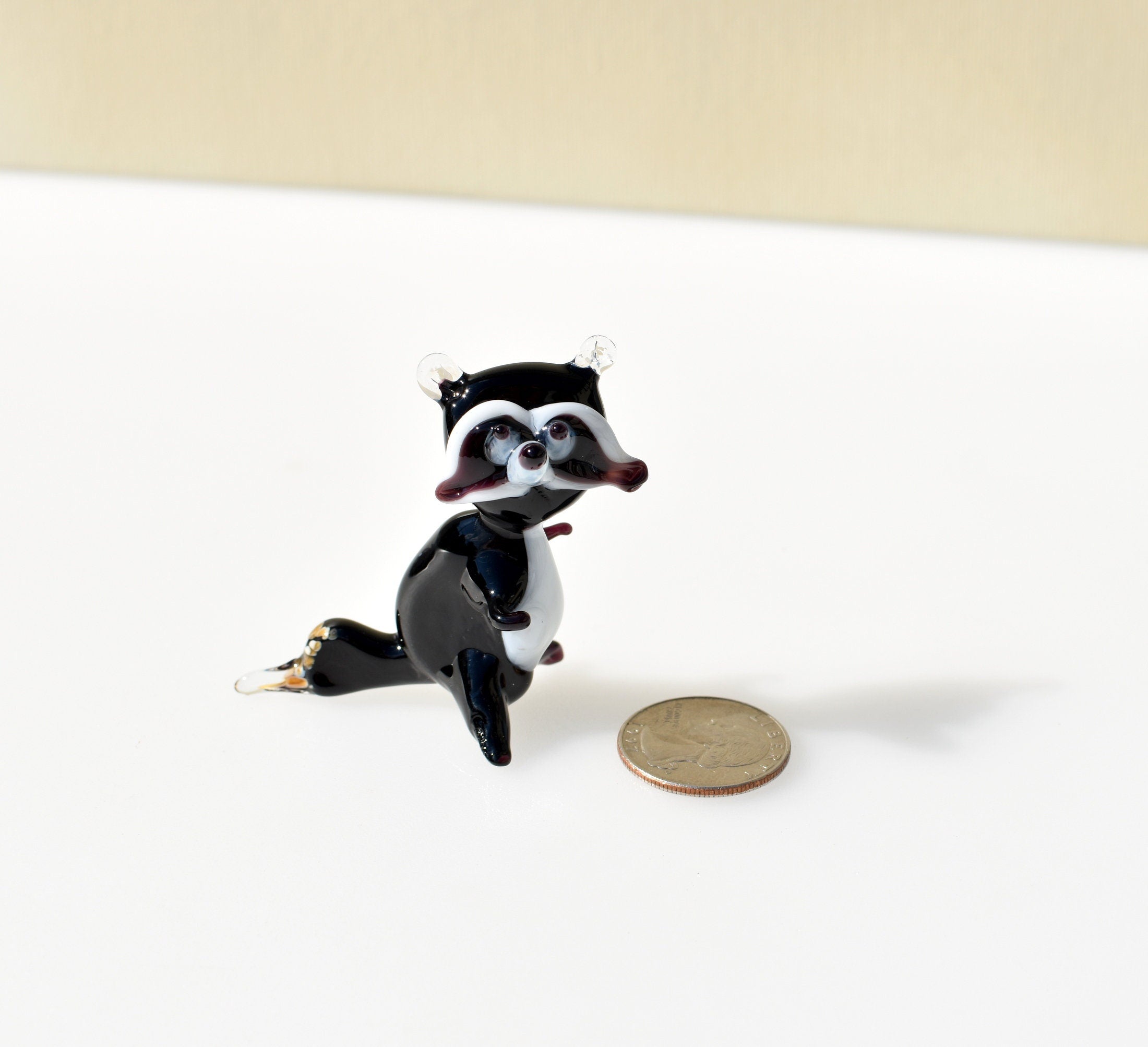 May include: A small glass figurine of a raccoon. The raccoon is black with white markings on its face and chest. The figurine is standing on a white surface with a silver coin in the background.