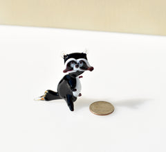 May include: A small glass figurine of a raccoon. The raccoon is black with white markings on its face and chest. The figurine is standing on a white surface with a silver coin in the background.
