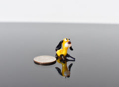 May include: A small yellow glass figurine of a dog with black ears and brown eyes. The dog is sitting on a black surface and is looking to the right. A silver quarter is in the foreground for scale.