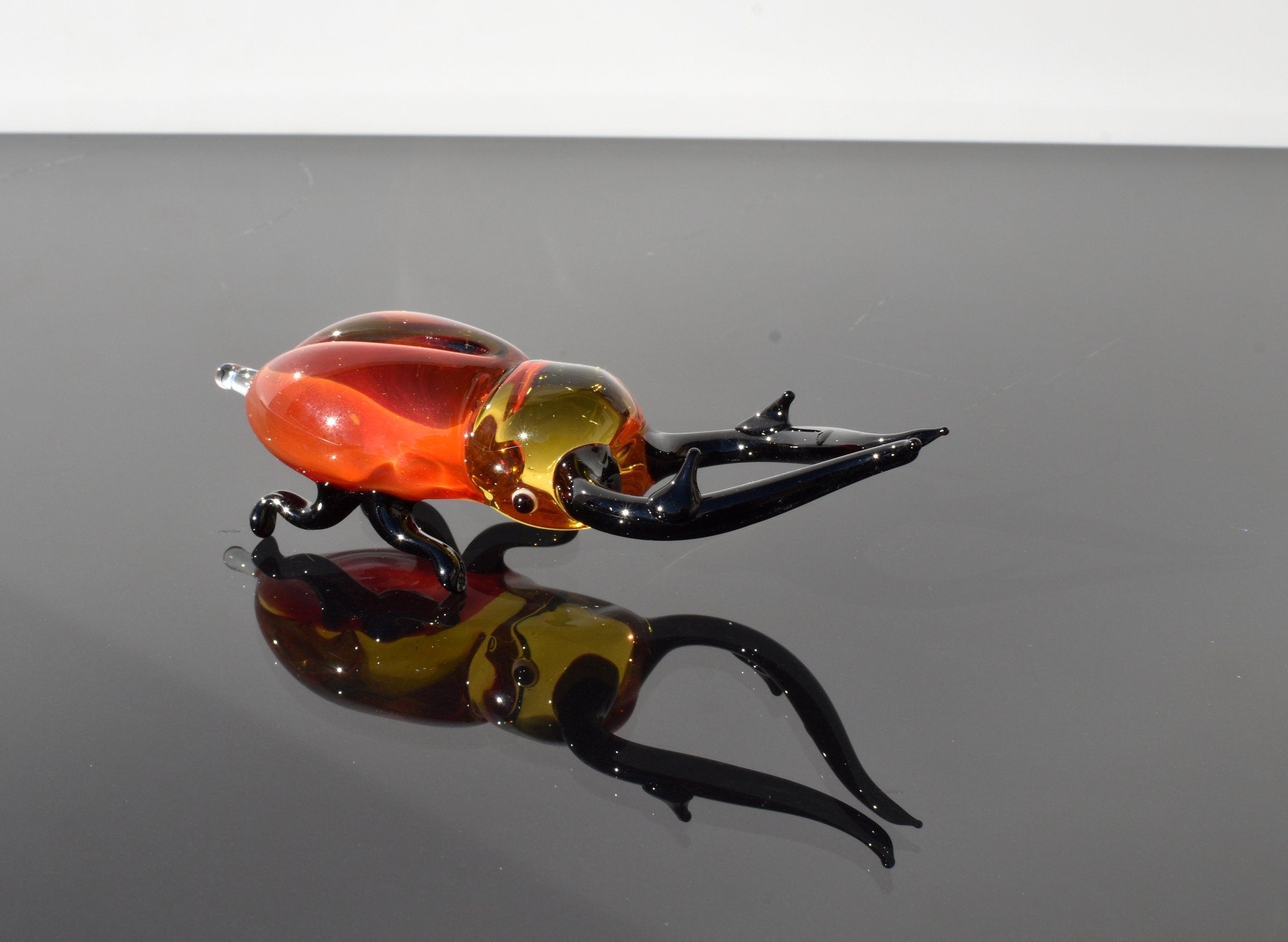 May include: A glass sculpture of a red and black stag beetle with long black antennae. The beetle is lying on its side with its legs outstretched. The sculpture is reflected in a black surface.