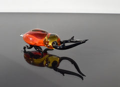 May include: A glass sculpture of a red and black stag beetle with long black antennae. The beetle is lying on its side with its legs outstretched. The sculpture is reflected in a black surface.