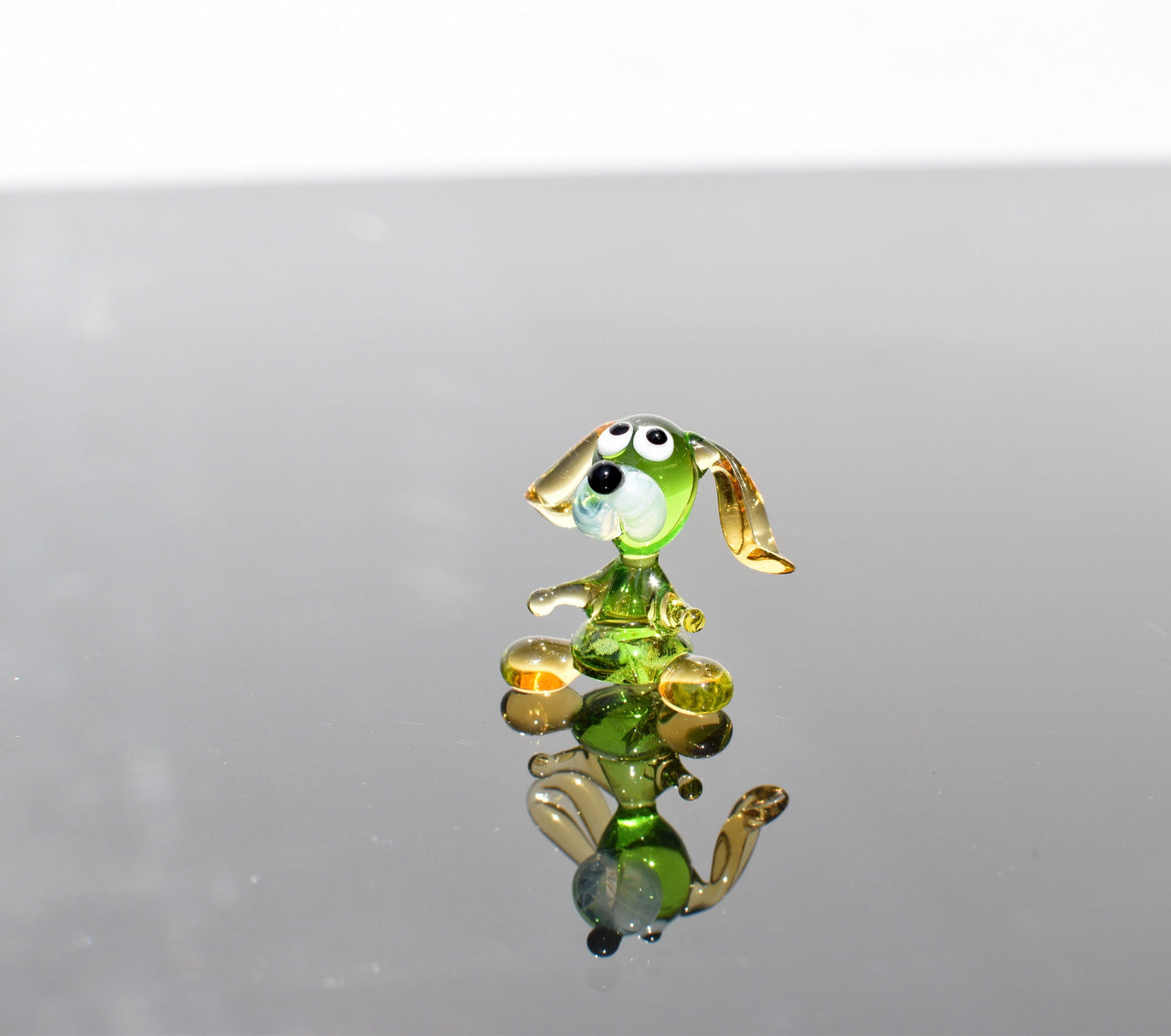May include: A small glass figurine of a dog. The dog is green with yellow ears and a white face. The dog is sitting on a reflective surface.