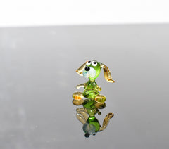 May include: A small glass figurine of a dog. The dog is green with yellow ears and a white face. The dog is sitting on a reflective surface.