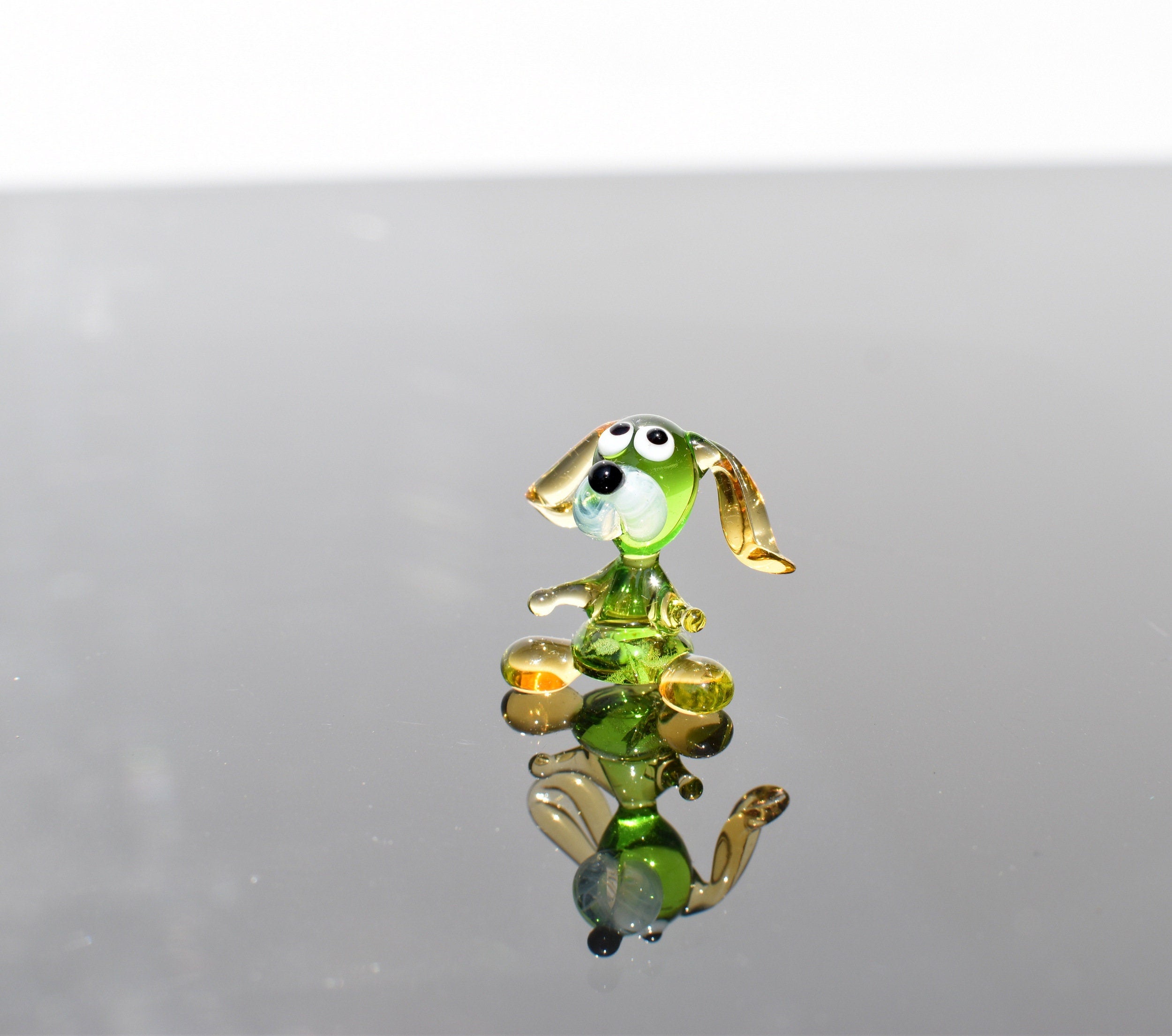 May include: A small glass figurine of a dog. The dog is green with yellow ears and a white face. The dog is sitting on a reflective surface.