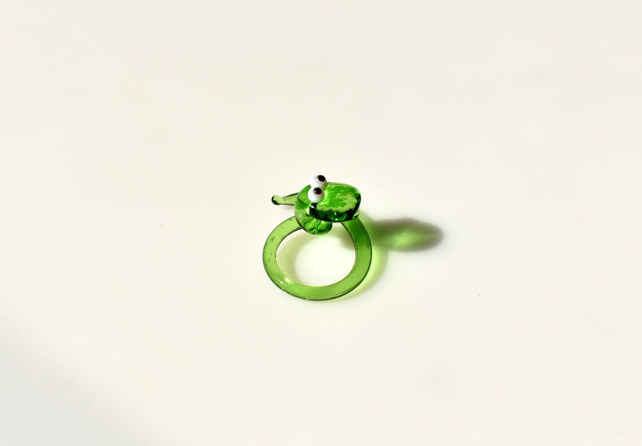 May include: A green glass snake figurine with white eyes. The snake is curled into a circle with its head facing forward. The snake is a decorative object.