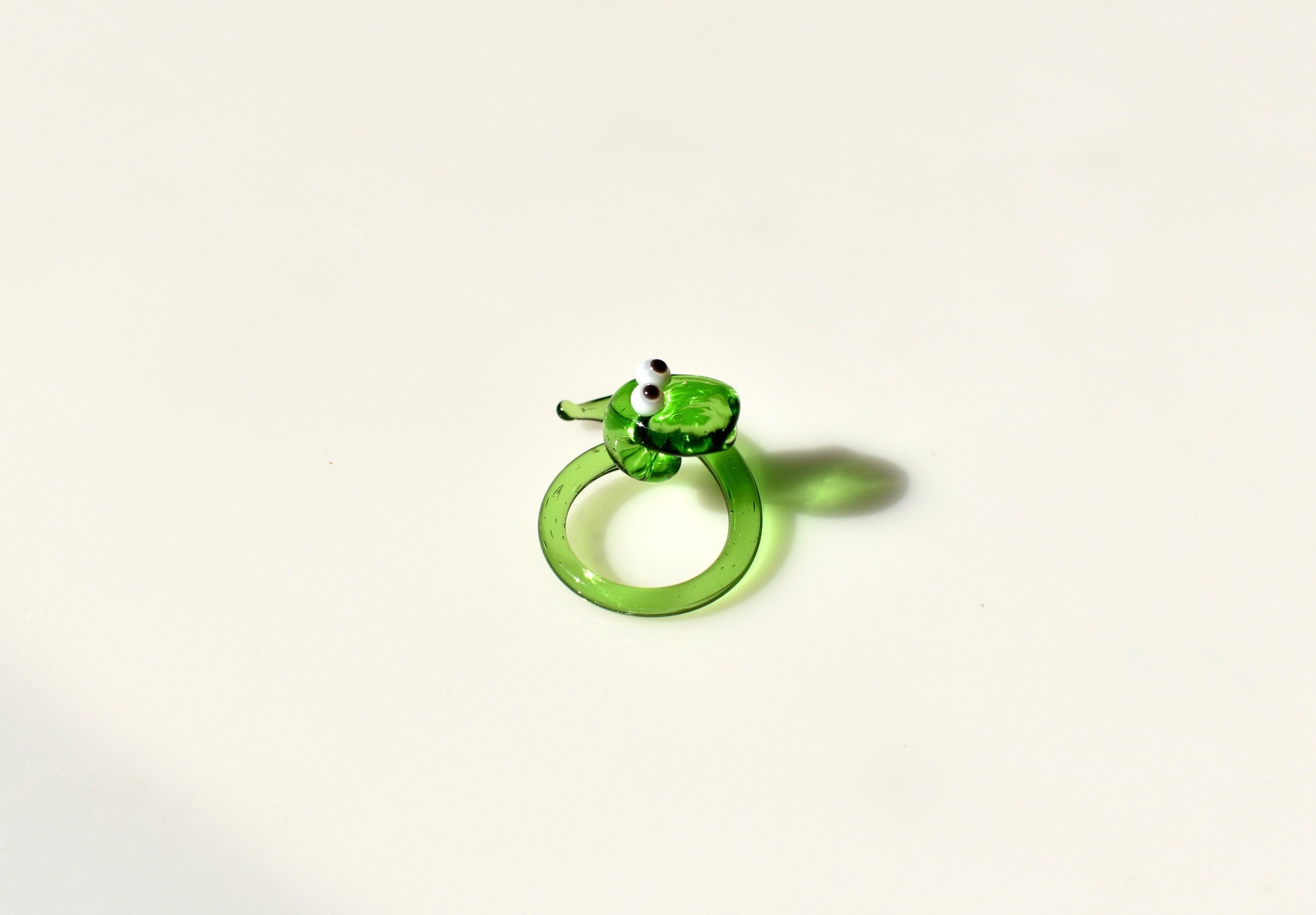 May include: A green glass snake figurine with white eyes. The snake is curled into a circle with its head facing forward. The snake is a decorative object.