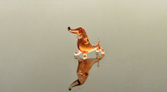 May include: A small glass dachshund figurine in a light brown color. The figurine is standing on a reflective surface and has a black nose.