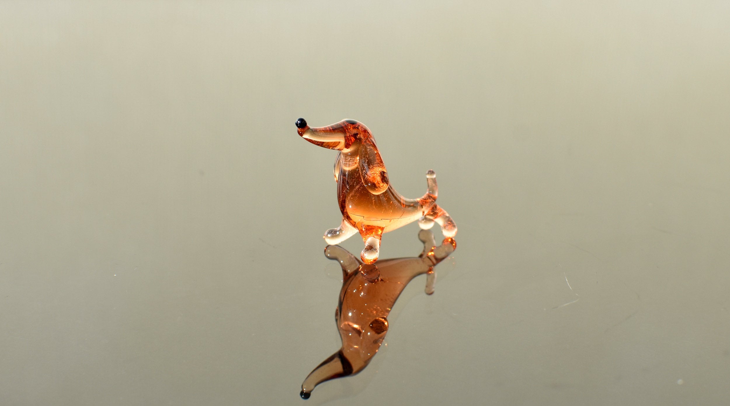 May include: A small glass dachshund figurine in a light brown color. The figurine is standing on a reflective surface and has a black nose.