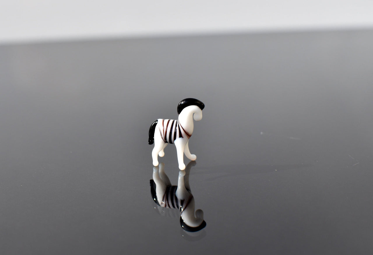 May include: A small glass zebra figurine with black and white stripes. The zebra is standing on a reflective surface and is facing to the right.