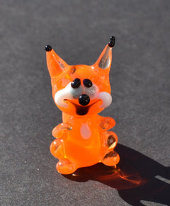 May include: A small orange glass figurine of a squirrel with black eyes and a white mustache. The squirrel is standing on a gray surface.