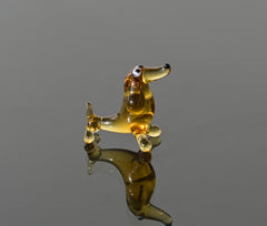 May include: A small glass dachshund figurine in amber color with black eyes and a black nose. The figurine is standing on a reflective surface.